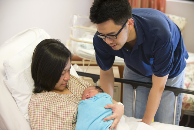 Mother lies in a hospital bed holding her newborn baby while father stands beside them and leans down to get a better view.