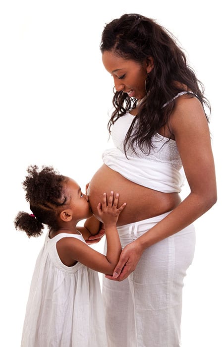Pregnant woman standing and her daughter standing in front of her and kissing her stomach.